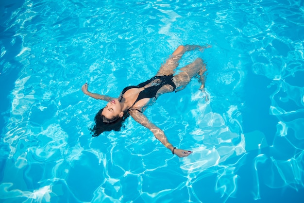 Attraktive frau in einem schwarzen badeanzug, der zurück auf sie in den swimmingpool schwimmt und sich entspannt Premium Fotos