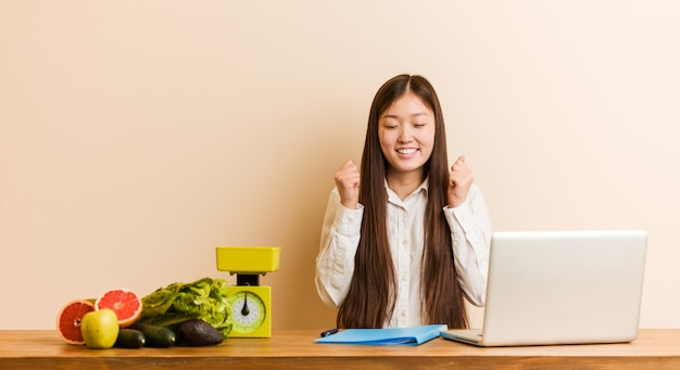 Chinesische frau des jungen ernährungswissenschaftlers, die mit ihrem laptop anhebt die faust arbeitet, glücklich und erfolgreich sich fühlt. Premium Fotos