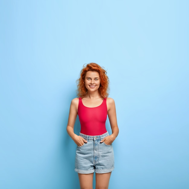 Das Vertikale Bild Der Jungen Frau Des Schlanken Ingwers Gekleidet In Der Roten Weste Und In Den Jeansshorts Halt Hande In Den Taschen Kostenlose Foto