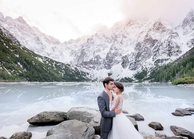 Das Zarte Verliebte Paar In Hochzeitsoutfits Umarmt Sich Vor Dem Zugefrorenen See Und Den Mit Schnee Bedeckten Hohen Bergen Kostenlose Foto