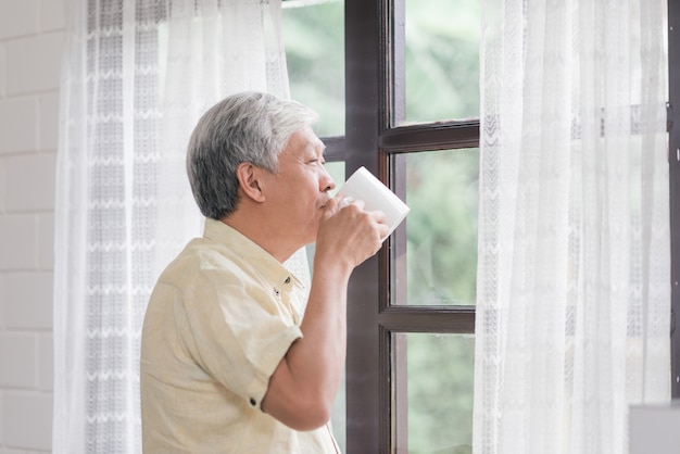 Der glückliche asiatische ältere mann, der einen tasse kaffee oder einen tee nahe dem fenster im wohnzimmer lächelt und trinkt, öffnen ältere asien-mann die vorhänge und entspannen sich morgens. Kostenlose Fotos