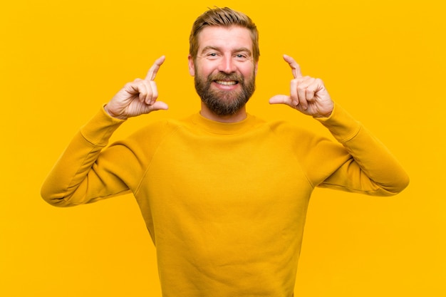 Der junge blonde mann, der eigenes lächeln mit beiden händen gestaltet oder umreißt, schaut positiv und glücklich, wellnessorangenwand Premium Fotos