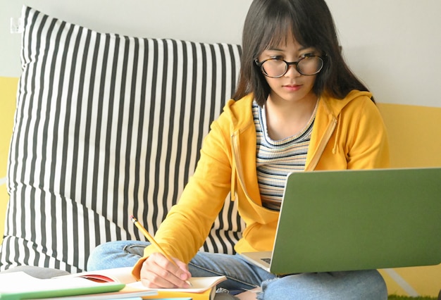Die asiatische studentin, die gläser trägt, erforscht mit einem laptop und nimmt kenntnisse, um einen bericht zu machen. Premium Fotos