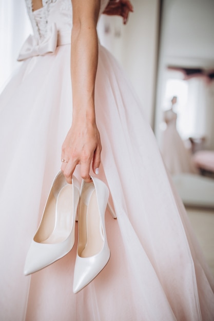 Die braut in ihrem hochzeitskleid hält ihre stilvollen beige schuhe in ihren händen. Premium Fotos