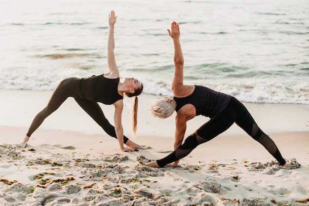 Die frauen, die eignung tun, trainieren in der synchronisierung, die in yoga asana auf dem strand nahe meer aufwirft Premium Fotos