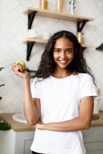 Die gelächelte mulattefrau, die im weißen t-shirt, mit hübschem gesicht und dem losen haar gekleidet wird, hält grünen apfel in der hand in der küche Kostenlose Fotos