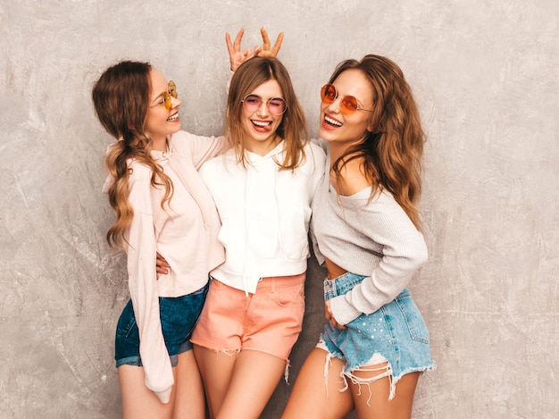Drei junge schöne lächelnde mädchen in der modischen sommersportkleidung. sexy sorglose frauenaufstellung. models in runden sonnenbrillen, die spaß haben. macht sich mit den fingern hörner auf den kopf Kostenlose Fotos