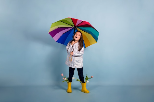 Ein Glückliches Kleines Mädchen In Einem Regenmantel Und Gelben Gummistiefeln Mit Blumen Hält 