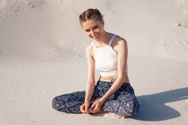 Ein schönes junges kaukasisches mädchen in einer weißen spitze und in einer weiten hose sitzt in einer lotussitz auf dem strand auf dem sand. die beliebteste pose für die meditation. lustig, lächelnd, glücklich Premium Fotos