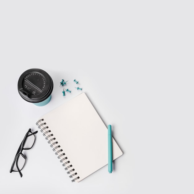 Eine draufsicht auf eine wegwerfbare kaffeetasse; stecknadeln; stift; brillen und spiralblock auf weißem hintergrund Kostenlose Fotos