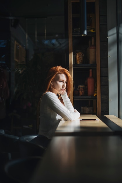 Eine einsame junge frau, die nahe dem fenster mit handy auf tabelle sitzt Kostenlose Fotos
