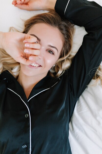 Entzuckendes Kaukasisches Madchen Im Schwarzen Pyjama Der Unter Decke Mit Lacheln Liegt Gluckseliges Weibliches Modell Mit Lockiger Frisur Die Sich Im Bett Entspannt Und Gesicht Mit Einer Hand Bedeckt Kostenlose Foto