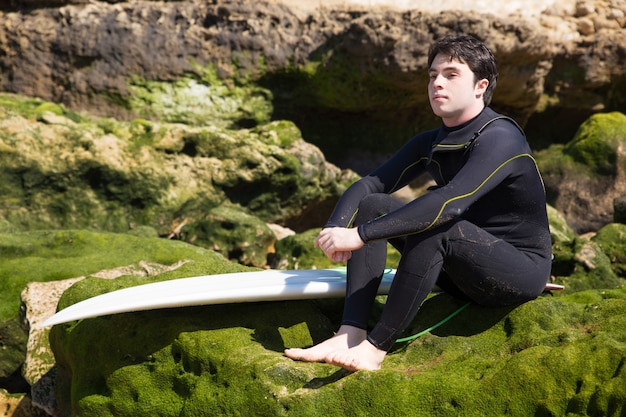 Ernster mann, der auf moosigen felsen mit surfbrett sitzt Kostenlose Fotos