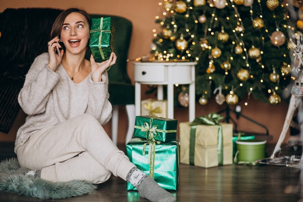 Frau, die durch weihnachtsbaum sitzt und am telefon kauft Kostenlose Fotos