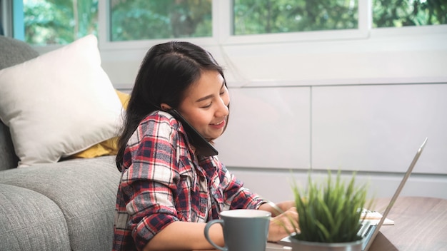 Freiberufliche asiatin, die zu hause arbeitet, die geschäftsfrau, die an laptop arbeitet und den handy zu hause spricht mit kunden auf sofa im wohnzimmer verwendet. Kostenlose Fotos