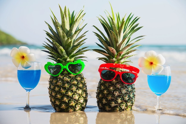 Frische paarananas mit sonnenbrillen und cocktailgläsern auf sauberem sand setzen mit seewelle - frische frucht und getränk mit meersandsonnen-ferienkonzept auf den strand Kostenlose Fotos