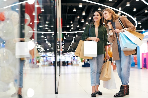 Frohliche Junge Frauen Mit Einkaufstuten Die An Der Vitrine Stehen Und Kleidung Im Einkaufszentrum Wahlen Premium Foto