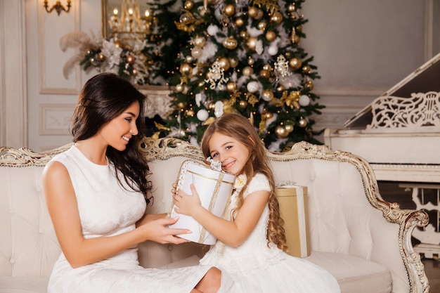 Frohe weihnachten und schöne feiertage. nette mutter und ihr nettes tochtermädchen, die geschenke im weißen klassischen innenklavier und in einem verzierten weihnachtsbaum austauscht. neujahr Premium Fotos