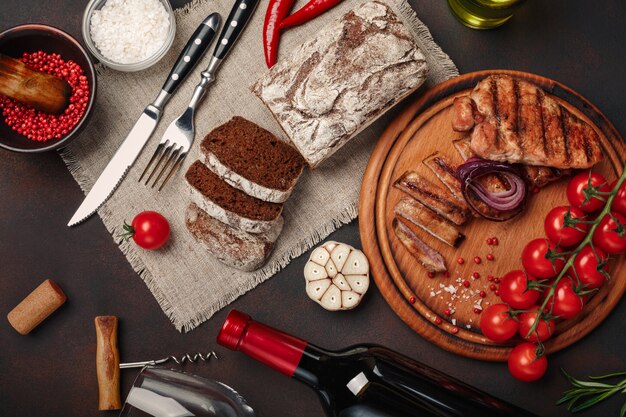 Gegrillte schweinefleischsteaks in scheiben geschnitten mit flasche wein, weinglas, korkenzieher, messer, gabel, schwarzbrot, kirschtomaten, knoblauch Premium Fotos