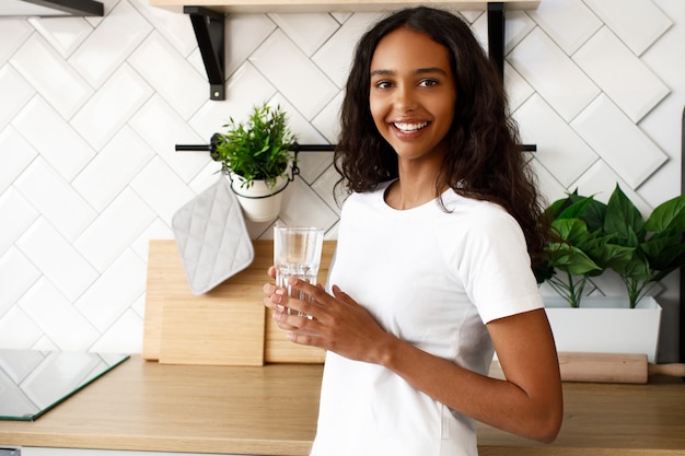 Gelächelte mulattefrau hält glas mit wasser nahe dem küchenschreibtisch