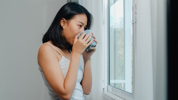 Glückliche schöne asiatin, die einen tasse kaffee oder einen tee nahe dem fenster im schlafzimmer lächelt und trinkt. offene vorhänge des jungen lateinischen mädchens und entspannen sich am morgen. lifestyle lady zu hause. Kostenlose Fotos