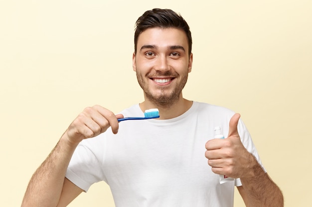 Glucklicher Energischer Junger Europaischer Kerl Mit Stoppeln Die Zahnburste Mit Bleichpaste Halten Und Daumen Hoch Geste Zeigen Die In Guter Stimmung Ist Kostenlose Foto