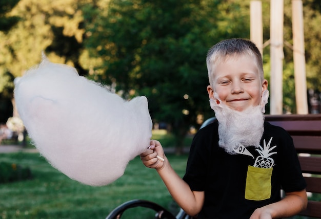 Glücklicher junge isst die zuckerwatte, die vom klebrigen gesponnenen zucker gemacht wird, der auf einer bank im park sitzt. happy boy machte einen bart aus zuckerwatte. Premium Fotos