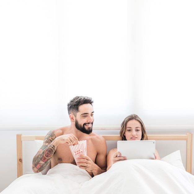 Glücklicher junger mann mit dem popcorn, das seine freundin verwendet digitale tablette auf bett betrachtet Kostenlose Fotos