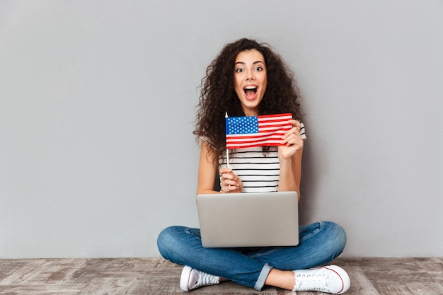 Herrliche frau mit dem schönen lächeln, das in der lotoshaltung mit silbernem computer auf den beinen demonstrieren amerikanische flagge über grauer wand sitzt Kostenlose Fotos