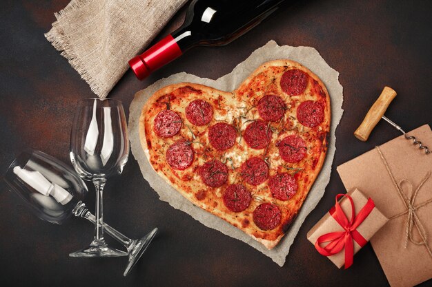 Herzförmige pizza mit mozzarella, wurst, weinflasche, weinglas zwei, geschenkbox auf rostigem hintergrund Premium Fotos