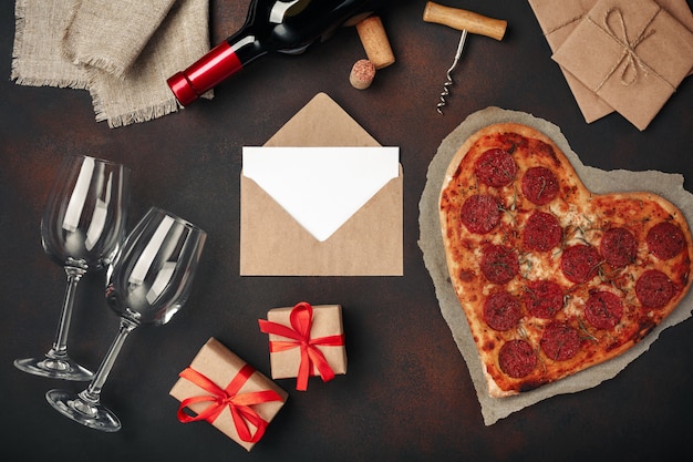 Herzförmige pizza mit mozzarella, wurst, weinflasche, weinglas zwei, geschenkbox auf rostigem hintergrund Premium Fotos