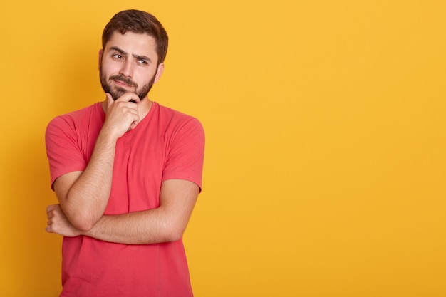 Horizontale Ernsthafte Unrasierte Mannliche Kleider Lassiges Rotes T Shirt Halt Die Hand Unter Dem Kinn Schaut Mit Ernstem Gesichtsausdruck Zur Seite Denkt An Etwas Posiert Auf Gelber Wand Mit Freiem Platz Kostenlose