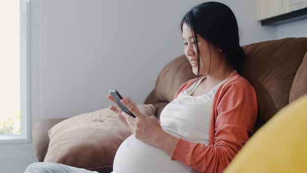 Junge asiatische schwangere frau, die handysuchschwangerschaftsinformationen verwendet. die mutter, die das glückliche lächeln positiv und ruhig sich fühlt, während mach s gut ihr kind, das zu hause auf sofa im wohnzimmer liegt. Kostenlose Fotos
