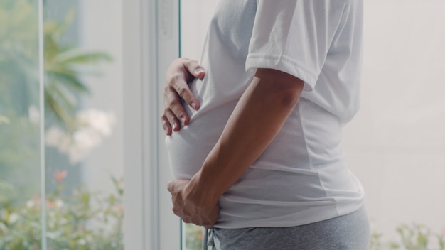 Junge asiatische schwangere frau, die ihren bauch spricht mit ihrem kind hält. die mutter, die sich glücklich fühlt, positiv und ruhig lächelnd, während mach s gut baby, schwangerschaft nahe fenster im wohnzimmer zu hause. Kostenlose Fotos