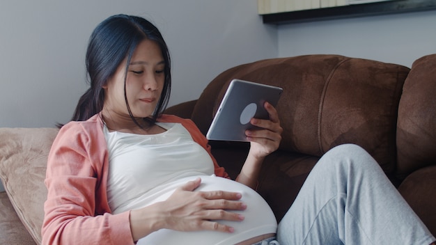 Junge asiatische schwangere frau, die tablettensuchschwangerschaftsinformationen verwendet. die mutter, die das glückliche lächeln positiv und ruhig sich fühlt, während mach s gut ihr kind, das zu hause auf sofa im wohnzimmer liegt. Kostenlose Fotos