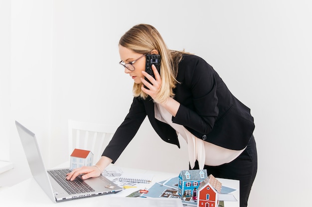 Junge attraktive frau, die am mobiltelefon beim arbeiten an laptop im immobilienbüro spricht Kostenlose Fotos