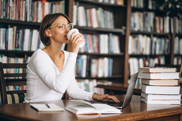Junge frau, die an der bibliothek unter verwendung der bücher und des computers sitzt Kostenlose Fotos