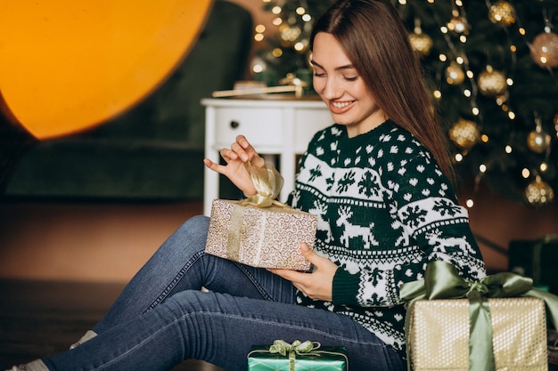Junge frau, die weihnachtsgeschenk durch den weihnachtsbaum auspackt Kostenlose Fotos
