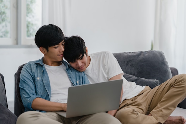 Junge homosexuelle paare unter verwendung des computerlaptops am modernen haus. das glückliche asiatische lgbtq + -männer entspannen sich spaß unter verwendung des aufpassenden films der technologie im internet zusammen, während lügensofa im wohnzimmer am haus. Kostenlose Fotos
