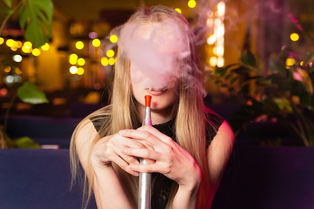 Junge kaukasische frau raucht eine huka oder eine shisha im nachtclub oder in der bar rauchen. nicht erkennbares gesicht hinter rauch Premium Fotos