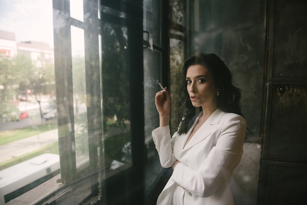 Junge sexy brunettefrau in der klage, die am fenster eine zigarette zuhause raucht Premium Fotos