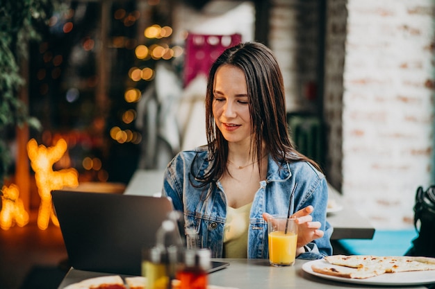 Junge studentin, die an laptop in der bar arbeitet und pizza isst Kostenlose Fotos