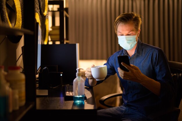 Junger Geschaftsmann Mit Maske Die Kaffee Halt Und Telefon Zu Hause Wahrend Der Arbeit Spat In Der Nacht Verwendet Premium Foto