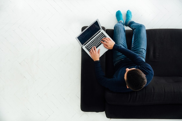 Junger mann, der an dem laptop, sitzend auf schwarzem sofa, weißer boden arbeitet. Kostenlose Fotos
