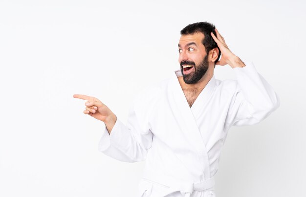 Junger mann, der karate über lokalisierter weißer wand überrascht tut und finger auf die seite zeigt Premium Fotos