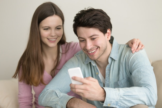 Junges glückliches paar, das zu hause auf sofa unter verwendung des smartphone sitzt Kostenlose Fotos