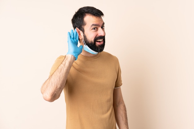 Kaukasischer Mann Mit Bart Der Mit Einer Maske Und Handschuhen Uber Isolierter Wand Schutzt Die Etwas Hort Indem Man Hand Auf Das Ohr Legt Premium Foto