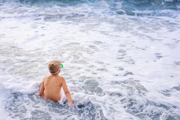 Kleiner netter kaukasischer kleinkindjunge mit dem blonden haar spielt am strand in den meereswellen am sonnigen sommertag. Premium Fotos