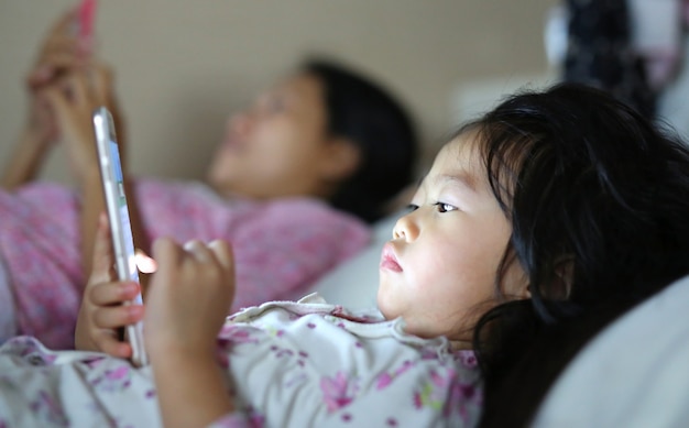 Kleines mädchen in den pyjamas mit der mutter, die den smartphone liegt auf einem bett spielt Premium Fotos