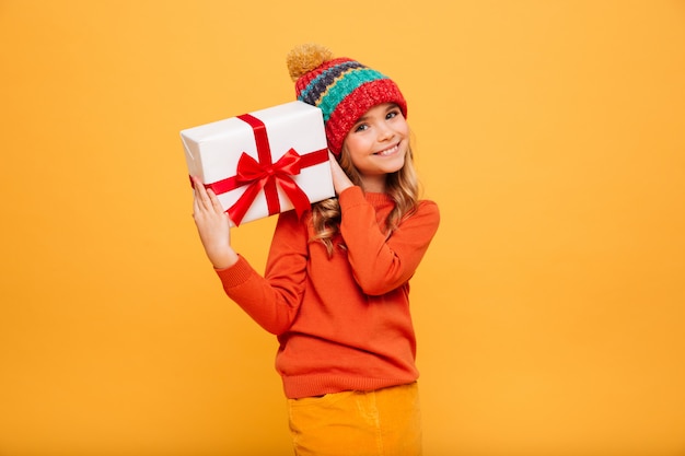 Lächelndes junges mädchen in der strickjacke und in hut, die geschenkbox halten und die kamera über orange betrachten Kostenlose Fotos
